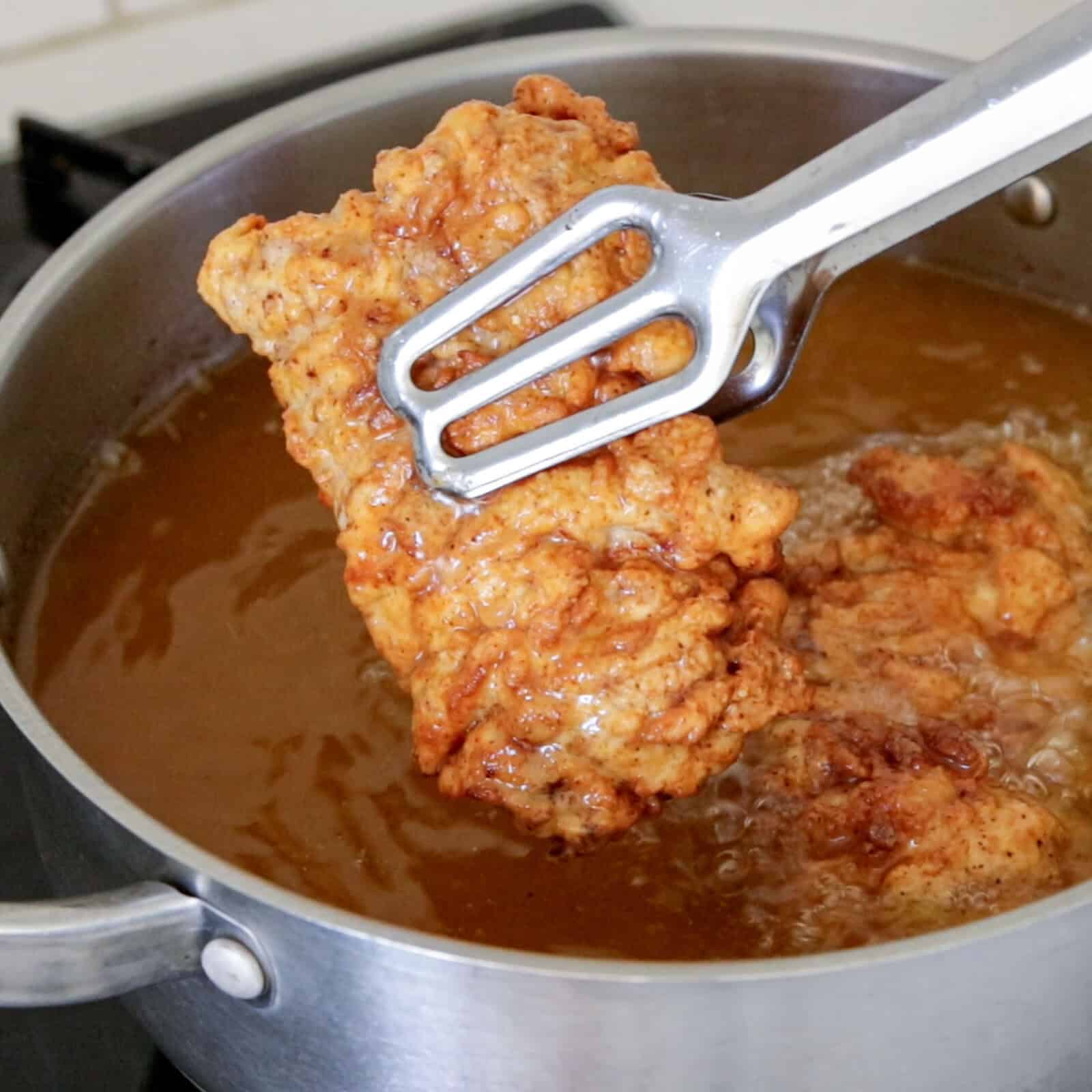 A piece of golden-brown fried chicken is being lifted out of hot oil in a stainless steel pot using metal tongs. The chicken is crispy and the oil is bubbling.