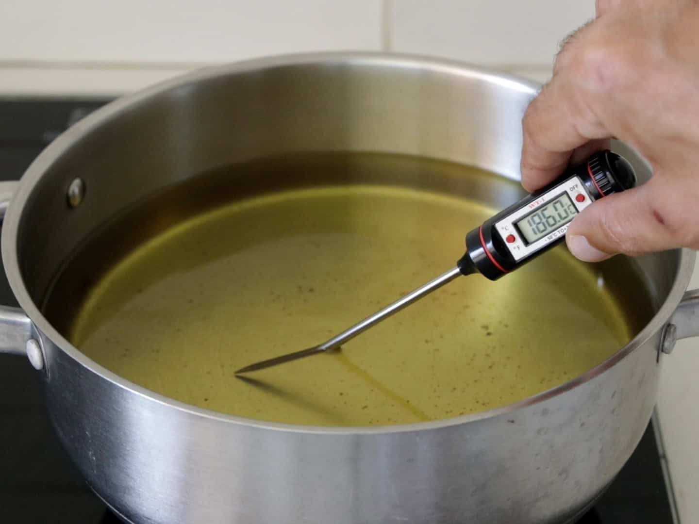 A hand holds a digital thermometer showing 186°C, checking the temperature of oil in a stainless steel pot on a stove.