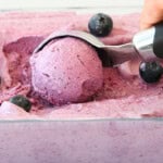 A hand uses an ice cream scoop to serve a ball of purple blueberry ice cream from a container, with whole blueberries scattered on top.