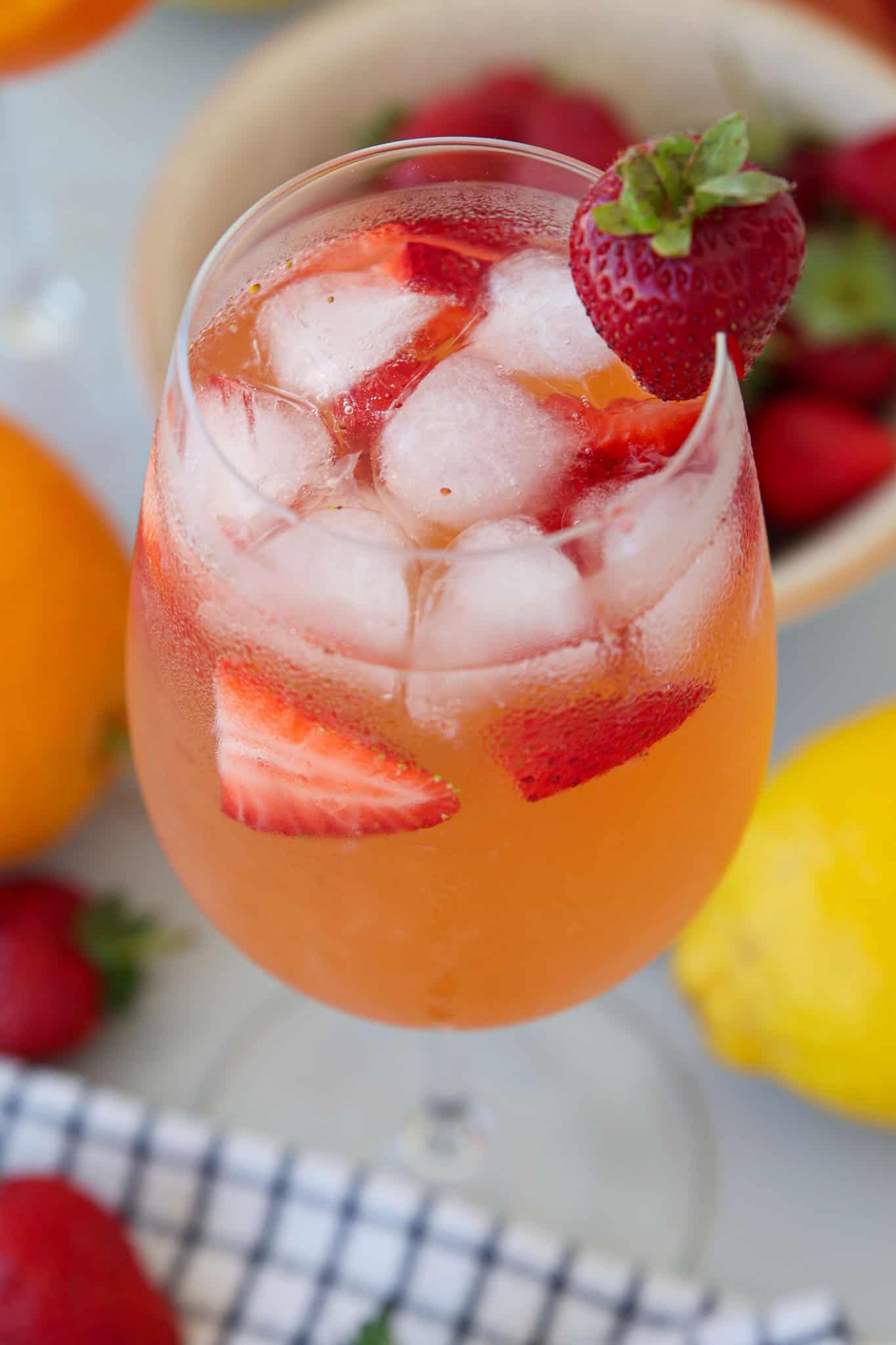 A glass of iced lemon strawberry sangria garnished with a strawberry on the rim, with sliced strawberries inside. Fresh strawberries, an orange, and a lemon are visible in the background.