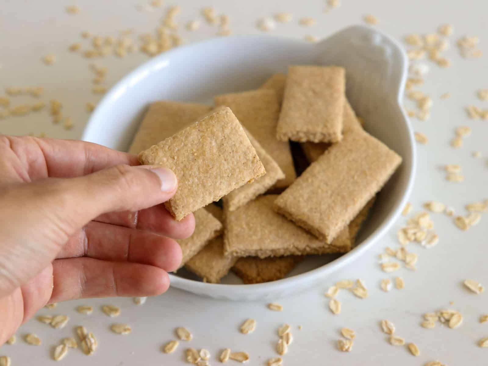 A hand holding a rectangular Gluten-Free Graham Crackers above a white bowl filled with similar crackers, with scattered oats on a light surface.