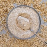 A bowl filled with oat flour and a spoon, surrounded by scattered whole rolled oats on a light surface.