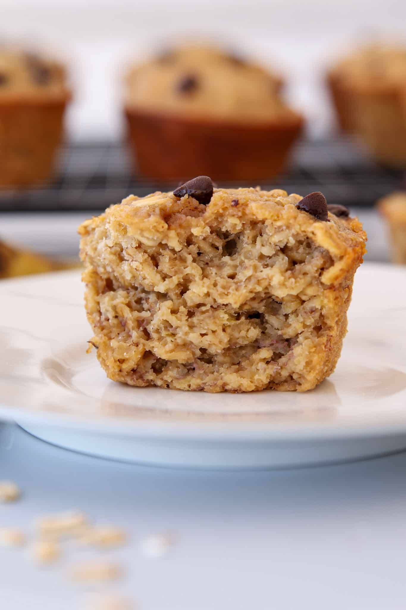 A close-up of a muffin with a moist, textured interior and chocolate chips on top, sitting on a white plate. More muffins are blurred in the background on a cooling rack.