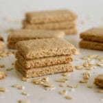 A close-up of stacked oat crackers on a white surface, with loose oats scattered around and more crackers blurred in the background.