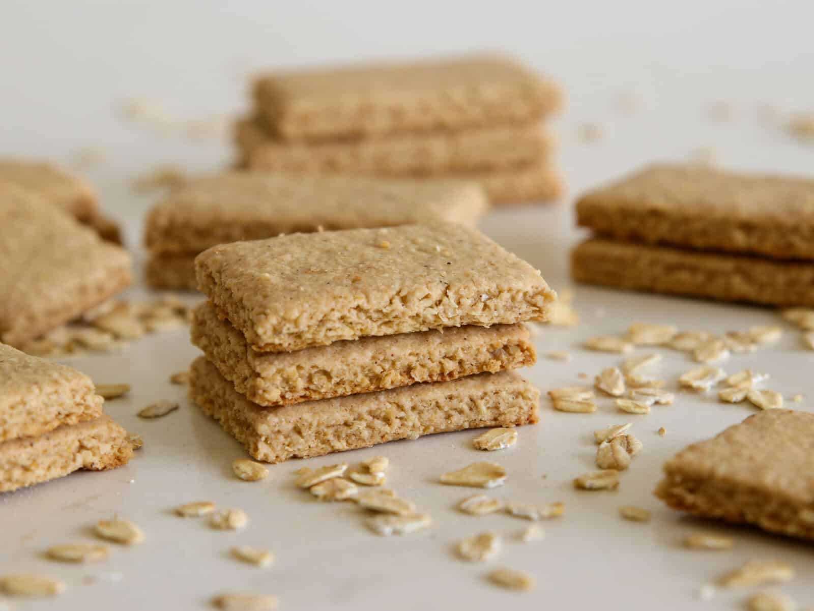A close-up of stacked oat crackers on a white surface, with loose oats scattered around and more crackers blurred in the background.