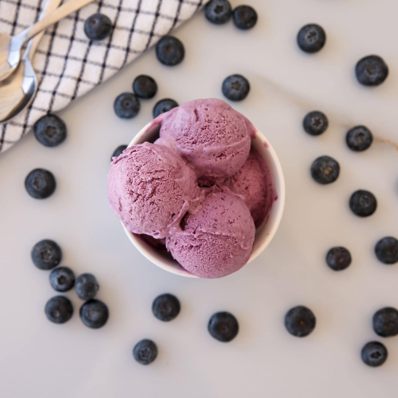 A bowl of three scoops of purple blueberry ice cream is surrounded by scattered fresh blueberries on a light surface, with a checkered cloth and two spoons nearby.