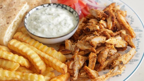 Juicy Chicken Gyros with Easy Greek Marinade A plate with crinkle-cut fries, spiced chicken shawarma, pita bread, sliced tomatoes, red onions, and a bowl of creamy tzatziki sauce.