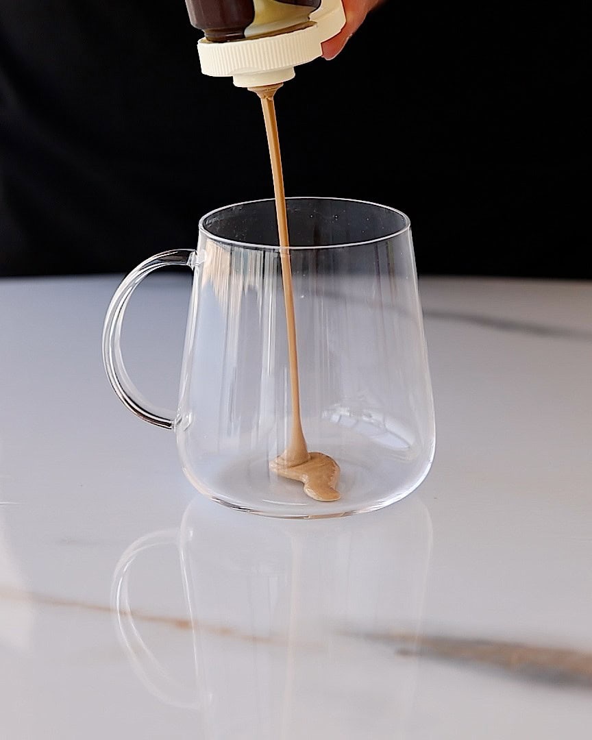 A hand squeezes a bottle, pouring a thick, light brown tahini into a clear glass mug on a white marble surface.