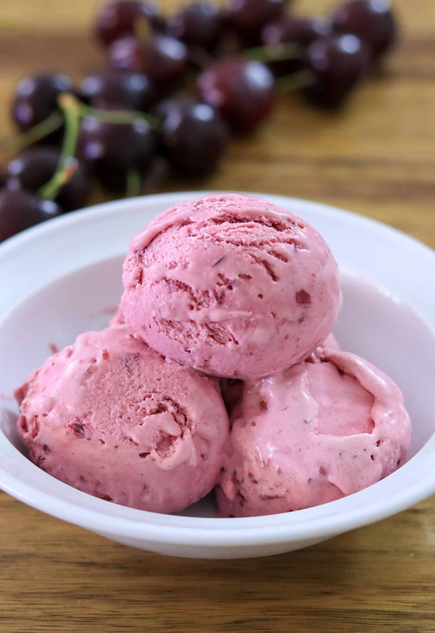 Three scoops of pink cherry ice cream in a white bowl on a wooden surface, with fresh cherries in the background.