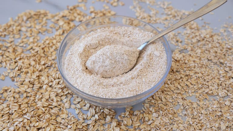 A glass bowl filled with oat flour has a spoon resting inside. Oat flakes are scattered around the bowl on a light surface.