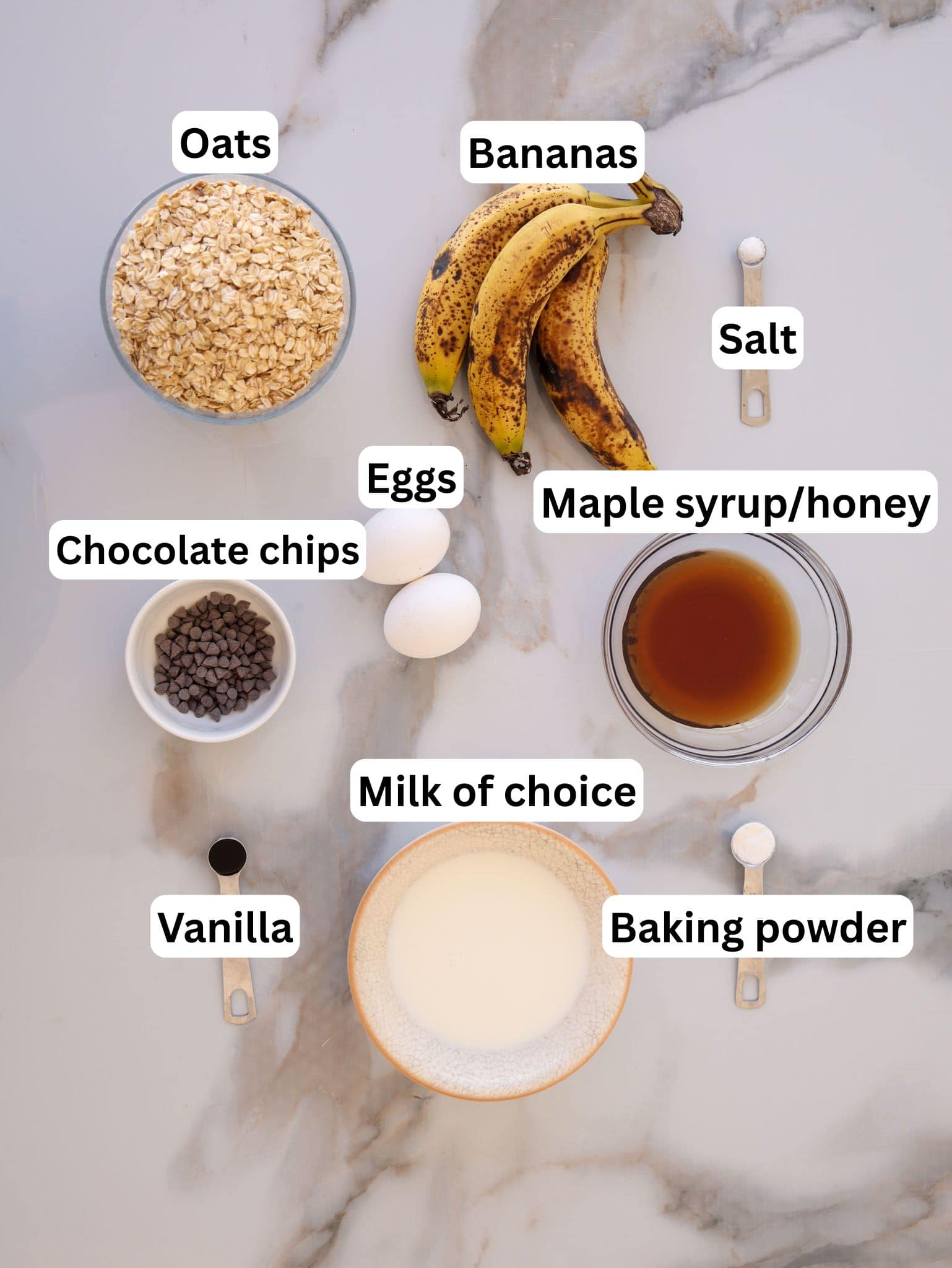 Overhead view of labeled ingredients on a marble surface: oats, bananas, eggs, salt, chocolate chips, maple syrup or honey, milk, vanilla, and baking powder.