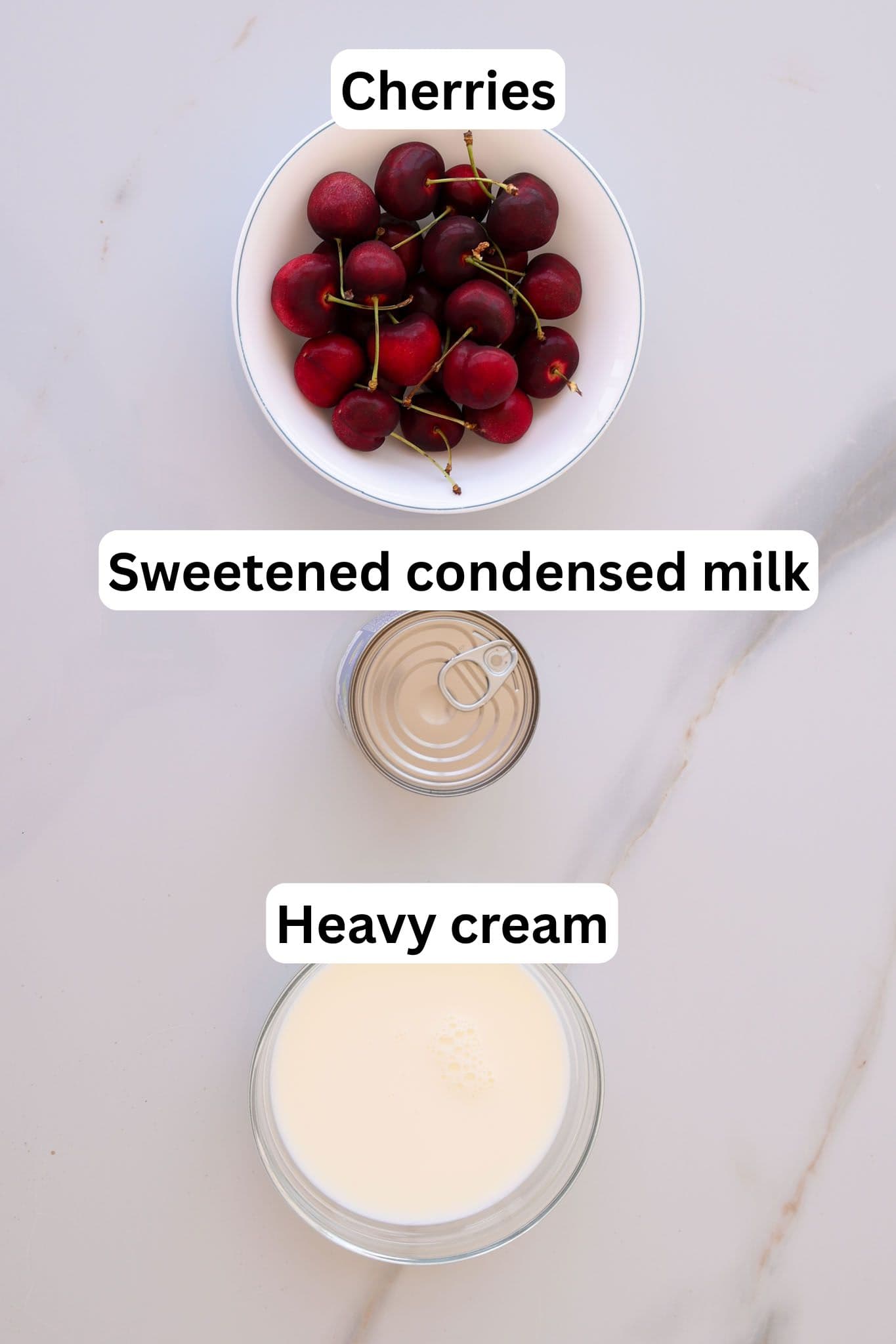 A bowl of cherries, an open can of sweetened condensed milk, and a bowl of heavy cream are arranged on a white surface, each labeled with text above: "Cherries," "Sweetened condensed milk," and "Heavy cream.
