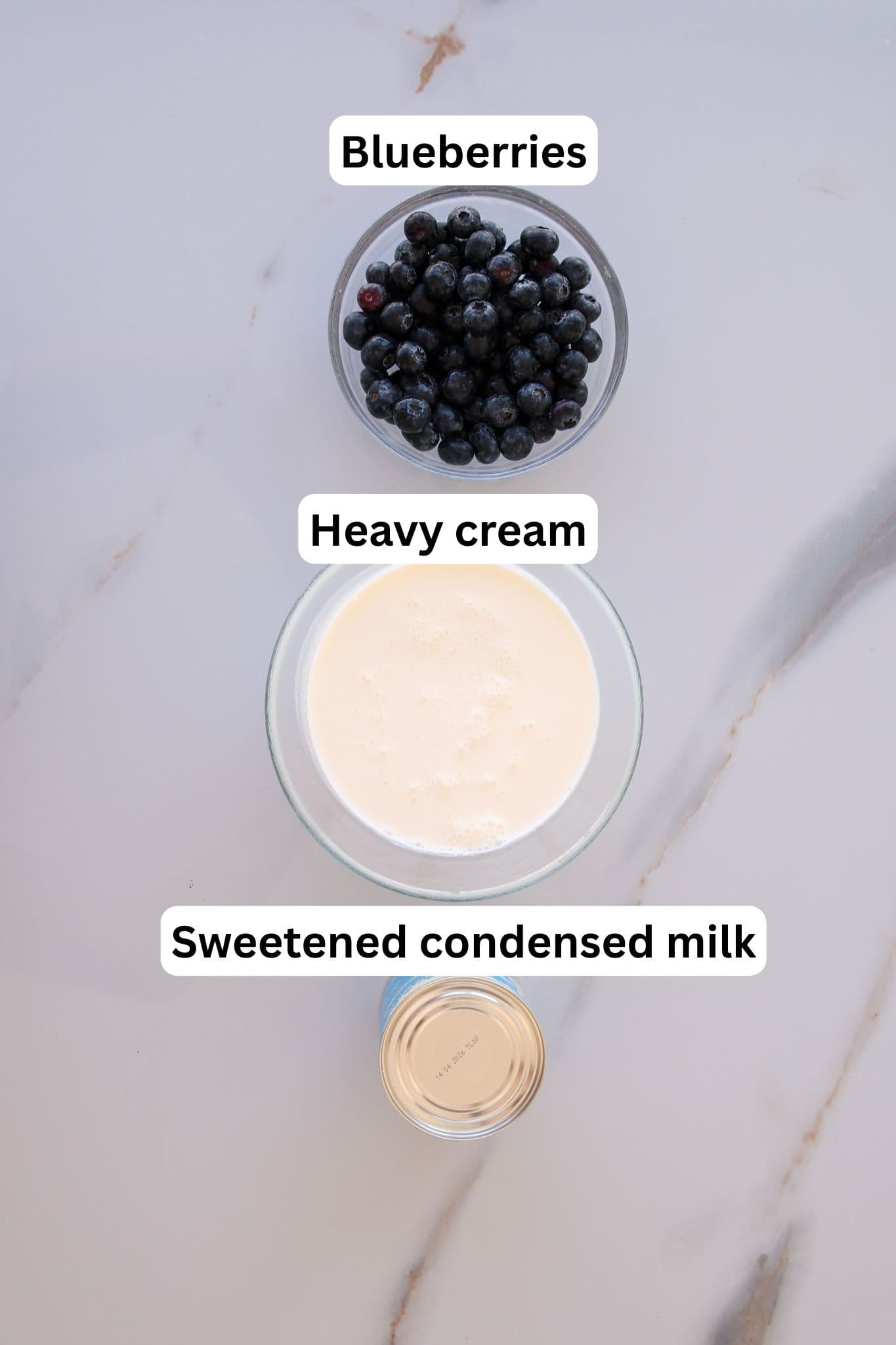 A flat lay photo of three ingredients on a marble surface: a bowl of blueberries at the top, a bowl of heavy cream in the middle, and an open can of sweetened condensed milk at the bottom, each labeled.
