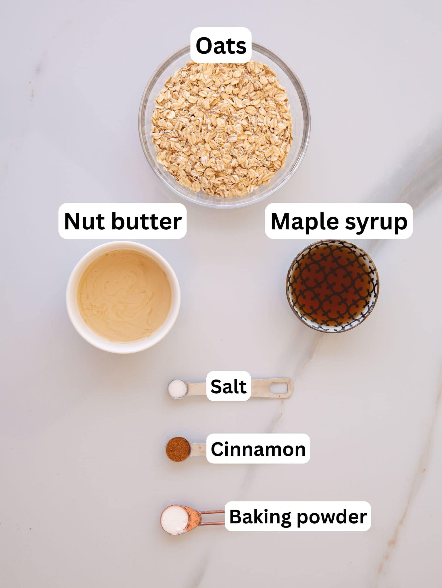 A flat lay of ingredients in bowls and spoons, labeled as: oats, nut butter, maple syrup, salt, cinnamon, and baking powder, arranged on a white marble surface.