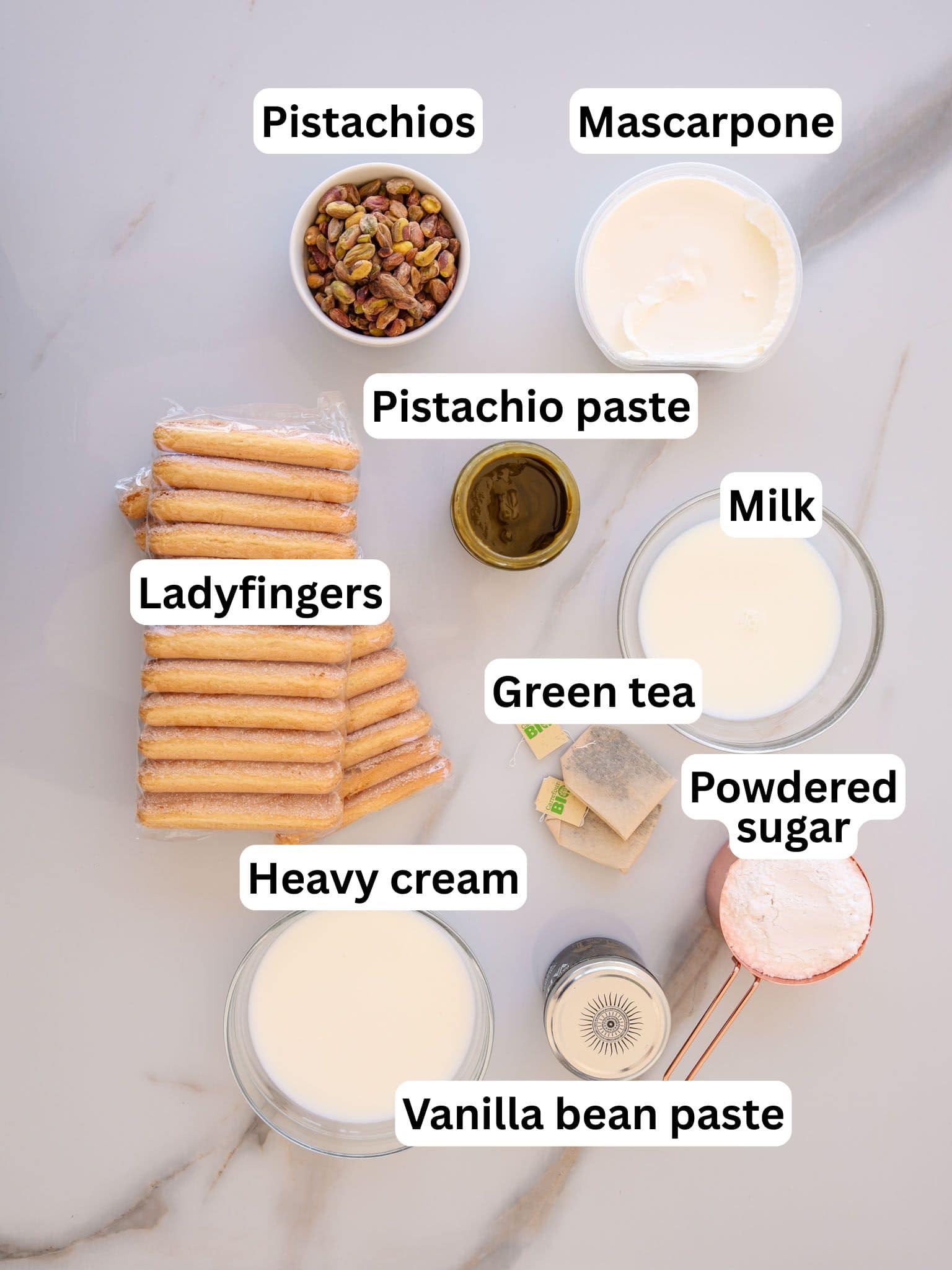 Ingredients for a dessert are arranged on a white surface, including bowls of pistachios, mascarpone, pistachio paste, milk, heavy cream, green tea bags, powdered sugar, vanilla bean paste, and a stack of ladyfingers.