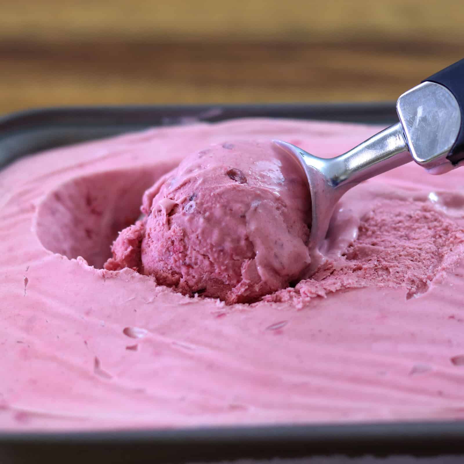 A metal ice cream scoop is lifting a scoop of pink ice cream from a container, with a smooth and creamy texture visible.
