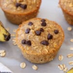 A banana oat muffin topped with chocolate chips sits on a light surface, surrounded by oats, a ripe banana, and other muffins in the background.