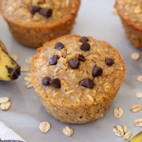 A banana oat muffin topped with chocolate chips sits on a light surface, surrounded by oats, a ripe banana, and other muffins in the background.