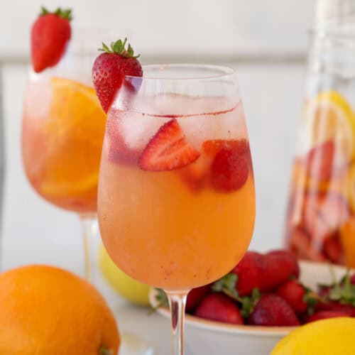 A chilled glass of fruity sangria garnished with strawberries, with sliced oranges, lemons, and strawberries around it. A pitcher filled with more fruit and sangria is in the background.