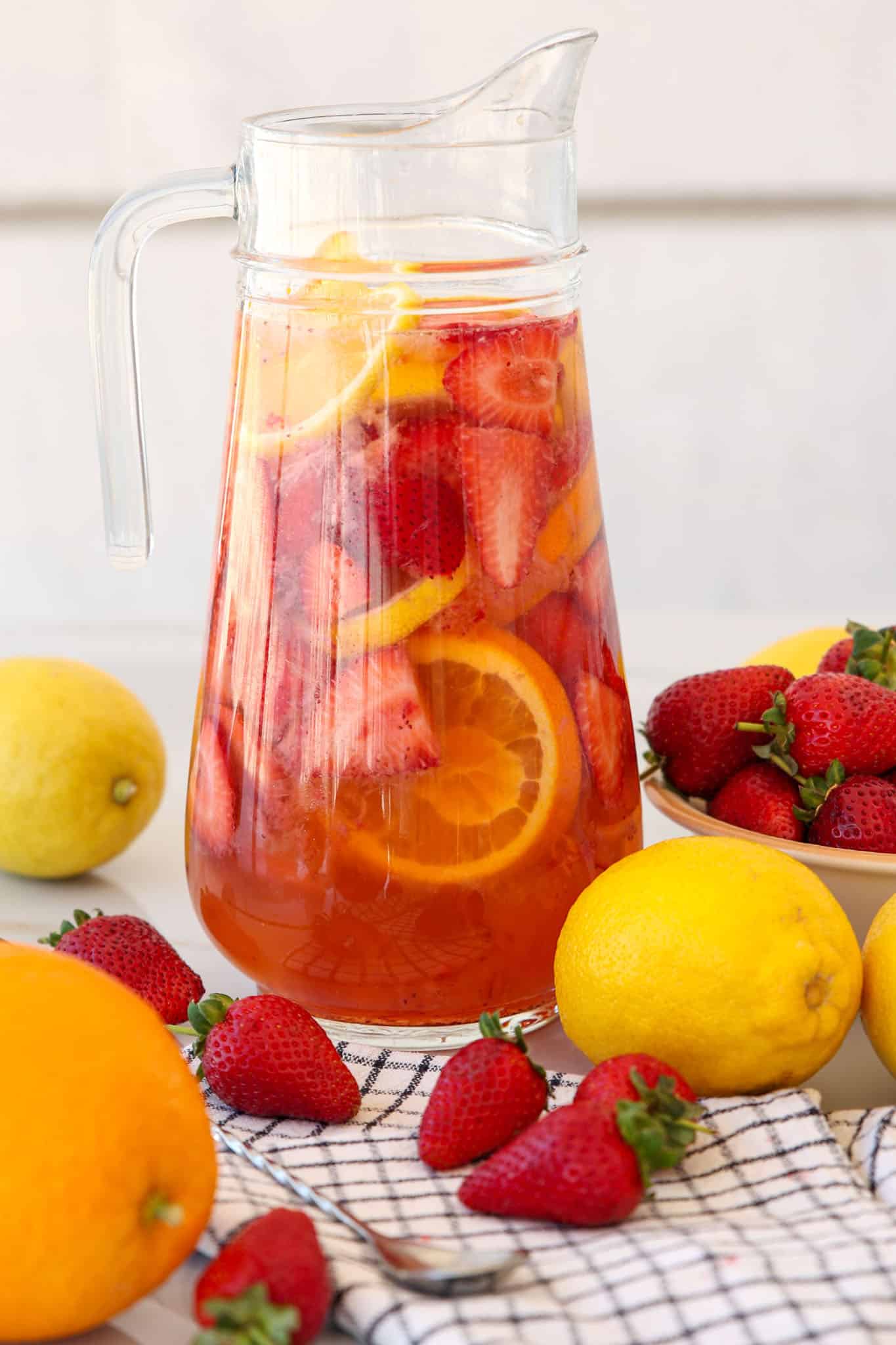 A glass pitcher filled with a refreshing strawberry sangria containing sliced strawberries, orange, and lemon sits on a table. Whole lemons, an orange, and strawberries are scattered nearby, along with a checkered cloth.