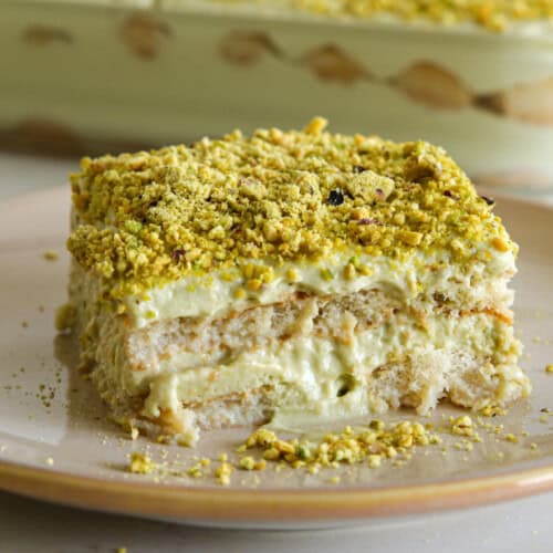 A square slice of creamy layered dessert topped with crushed pistachios sits on a beige plate, with the remaining dessert visible in the background.