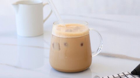 The Viral Tahini Coffee That Broke the Internet A glass mug of iced tahini coffee with a clear straw sits on a white marble surface. There are visible ice cubes in the drink, and a white ceramic pitcher is blurred in the background.