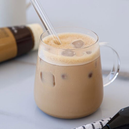 A glass cup filled with iced coffee and a clear straw sits on a light surface. There is a plaid cloth and a bottle of coffee creamer nearby, with a white mug in the background.
