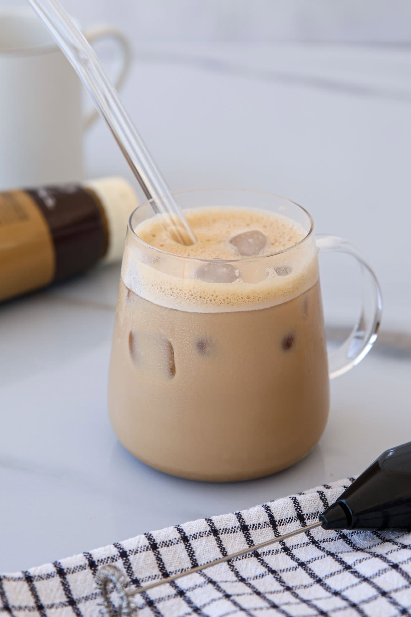 A glass cup filled with iced coffee with tahini and a clear straw sits on a light surface. There is a plaid cloth and a bottle of coffee creamer nearby, with a white mug in the background.