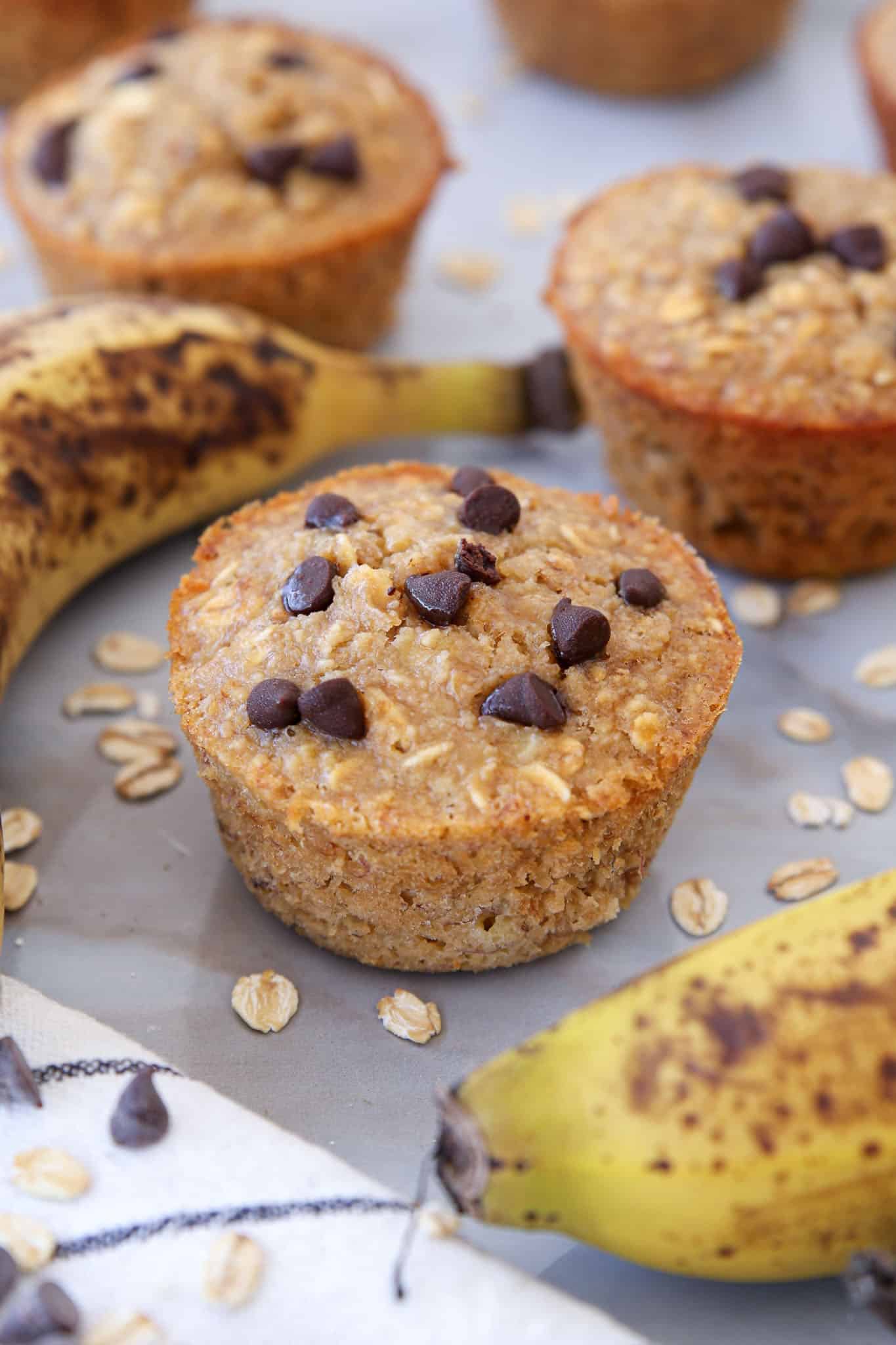 Banana oatmeal muffins topped with chocolate chips are surrounded by ripe, spotty bananas and scattered oats on a light surface. Some muffins are in focus while others are blurred in the background.