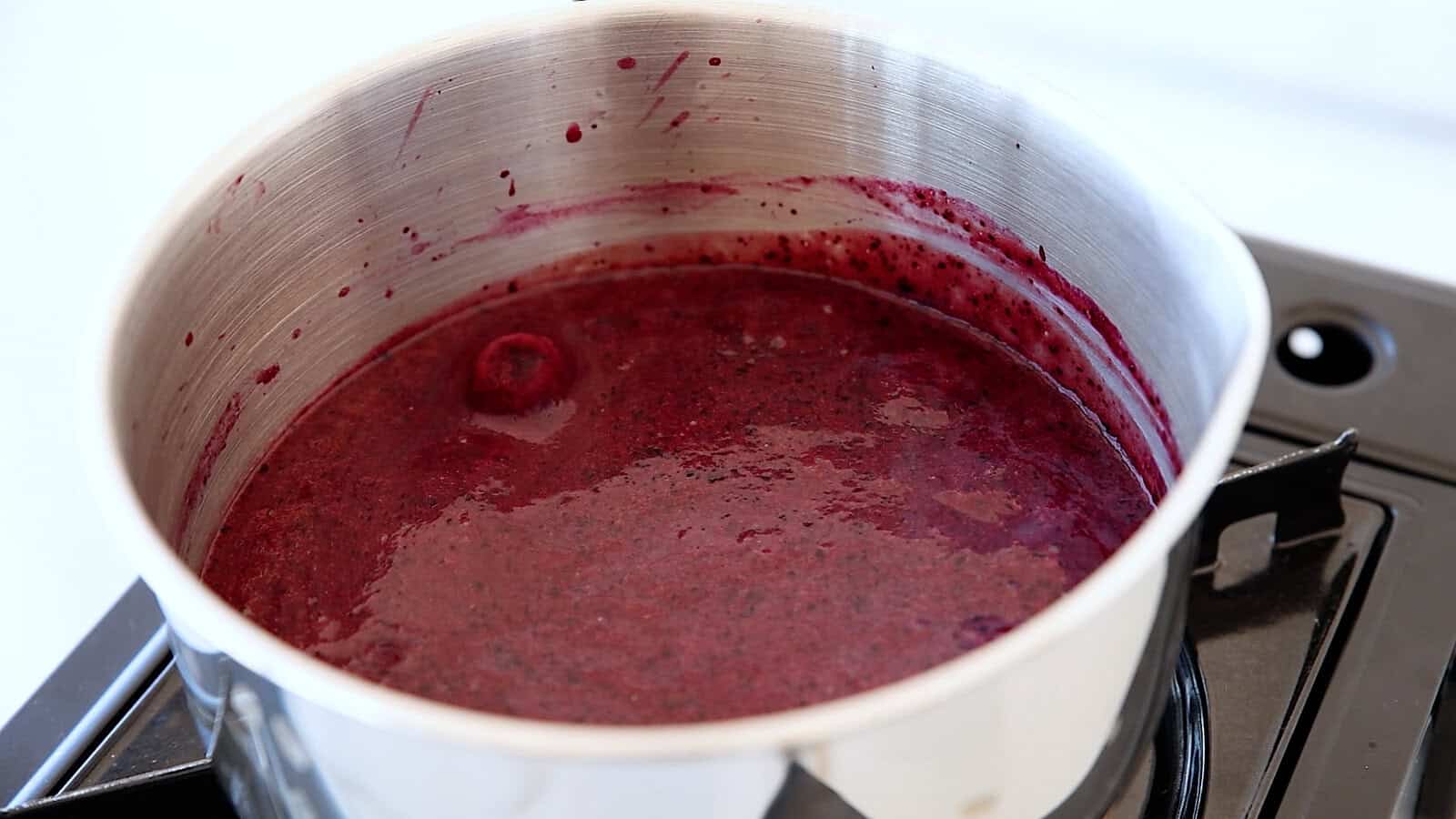 A stainless steel pot on a stove contains a thick, dark purple mixture, likely a fruit sauce or jam, simmering with bubbles and splashes visible along the sides of the pot.