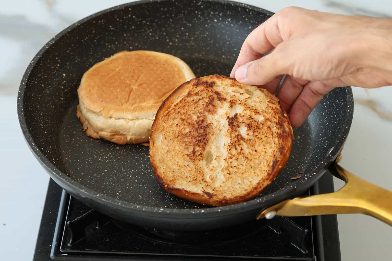 A hand lifts a toasted hamburger bun from a black frying pan while another bun half toasts next to it on a stovetop.