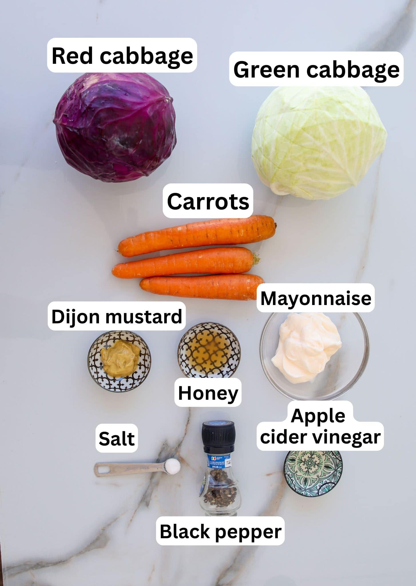 Overhead view of ingredients on a white surface: a red cabbage, a green cabbage, three carrots, bowls of mayonnaise, Dijon mustard, honey, apple cider vinegar, salt in a spoon, and a black pepper grinder. Labels identify each item.
