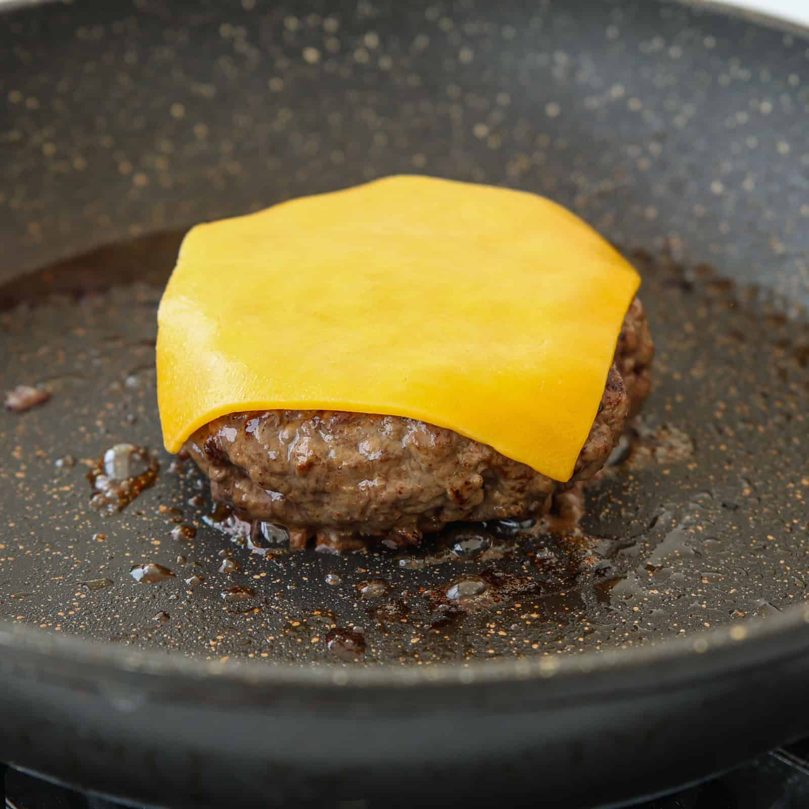A beef patty topped with a slice of melted cheddar cheese is cooking in a black skillet.