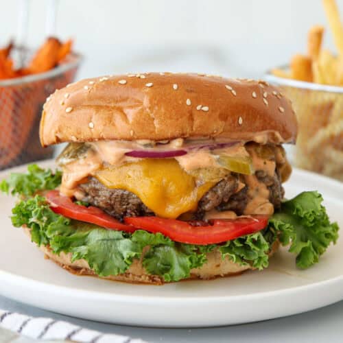 A cheeseburger with lettuce, tomato, pickles, onions, cheddar cheese, and sauce on a sesame bun sits on a white plate, with baskets of sweet potato fries and regular fries in the background.
