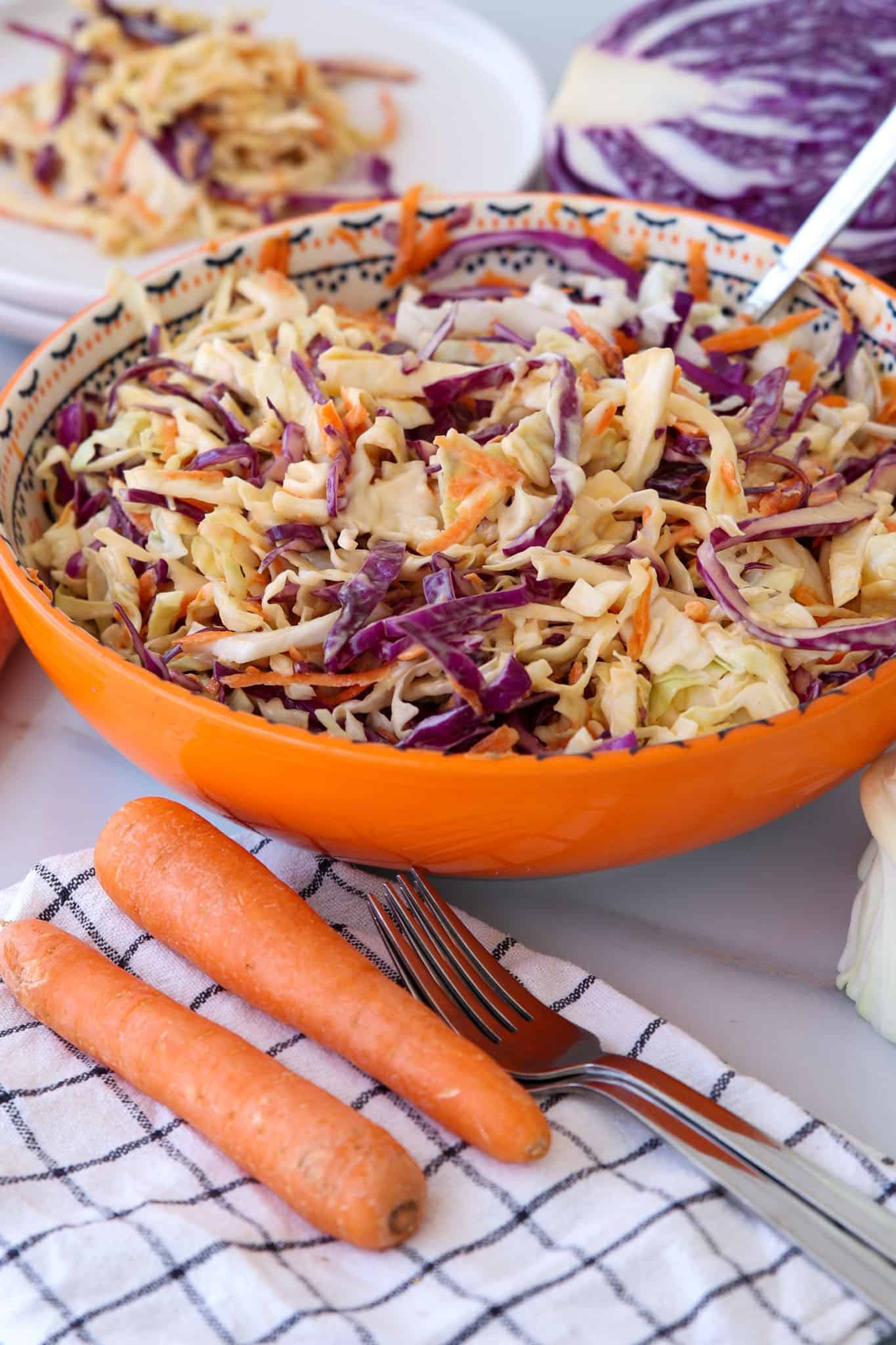 A bowl of coleslaw with shredded green and purple cabbage and carrots sits on a table next to two whole carrots, a fork, and a checkered napkin. Sliced cabbage and more coleslaw are in the background.