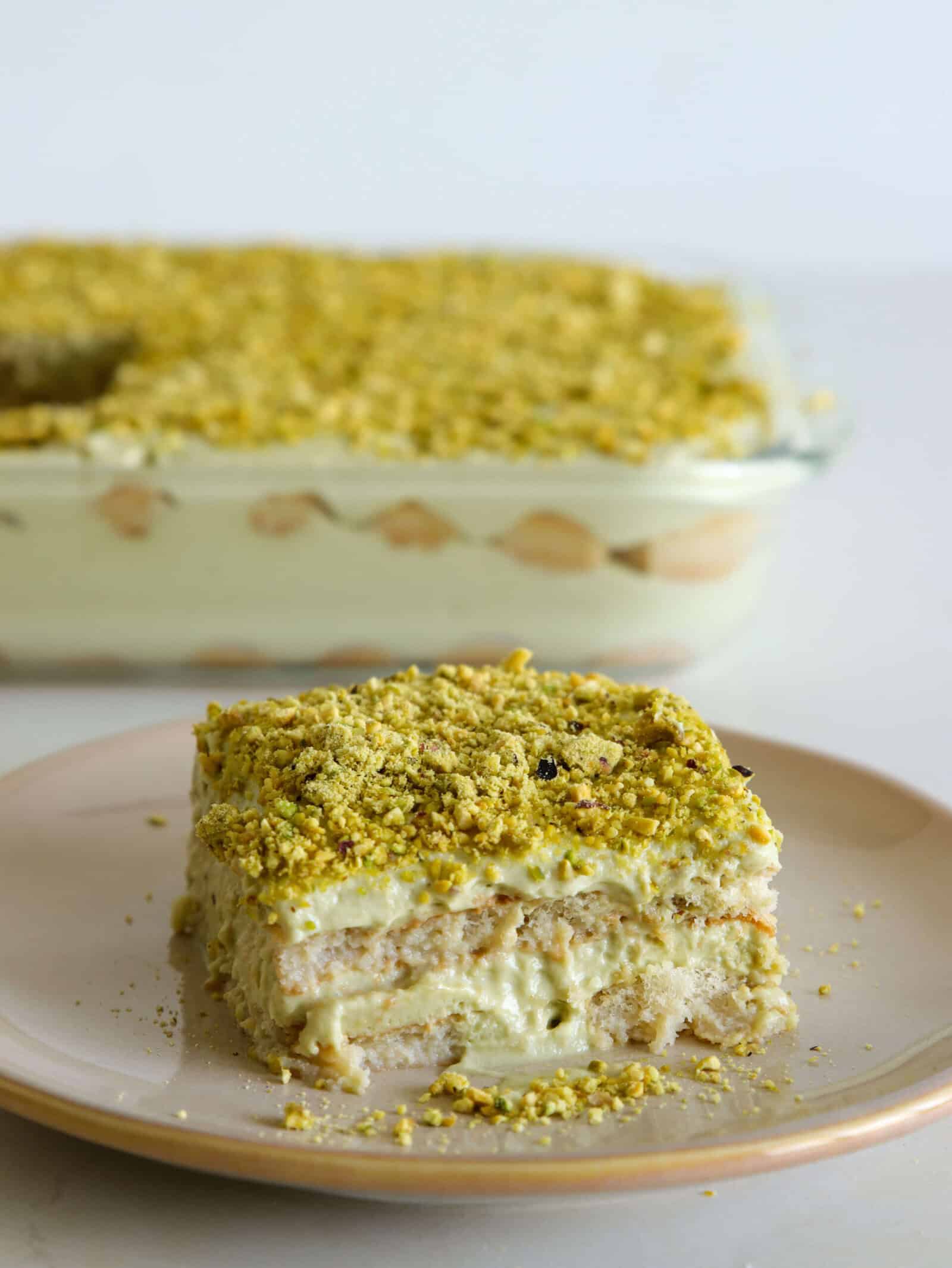 A square slice of creamy pistachio tiramisu topped with crushed pistachios sits on a plate, with the rest of the dessert in a glass dish in the background; The dessert has visible layers of cream and cake or biscuit.