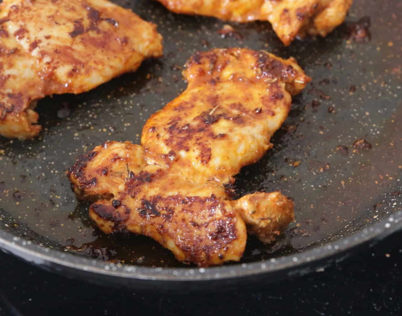 Seasoned chicken breasts are cooking in a black frying pan. The chicken pieces are browned with visible spices and slight charring, and there is some oil and seasoning residue in the pan.