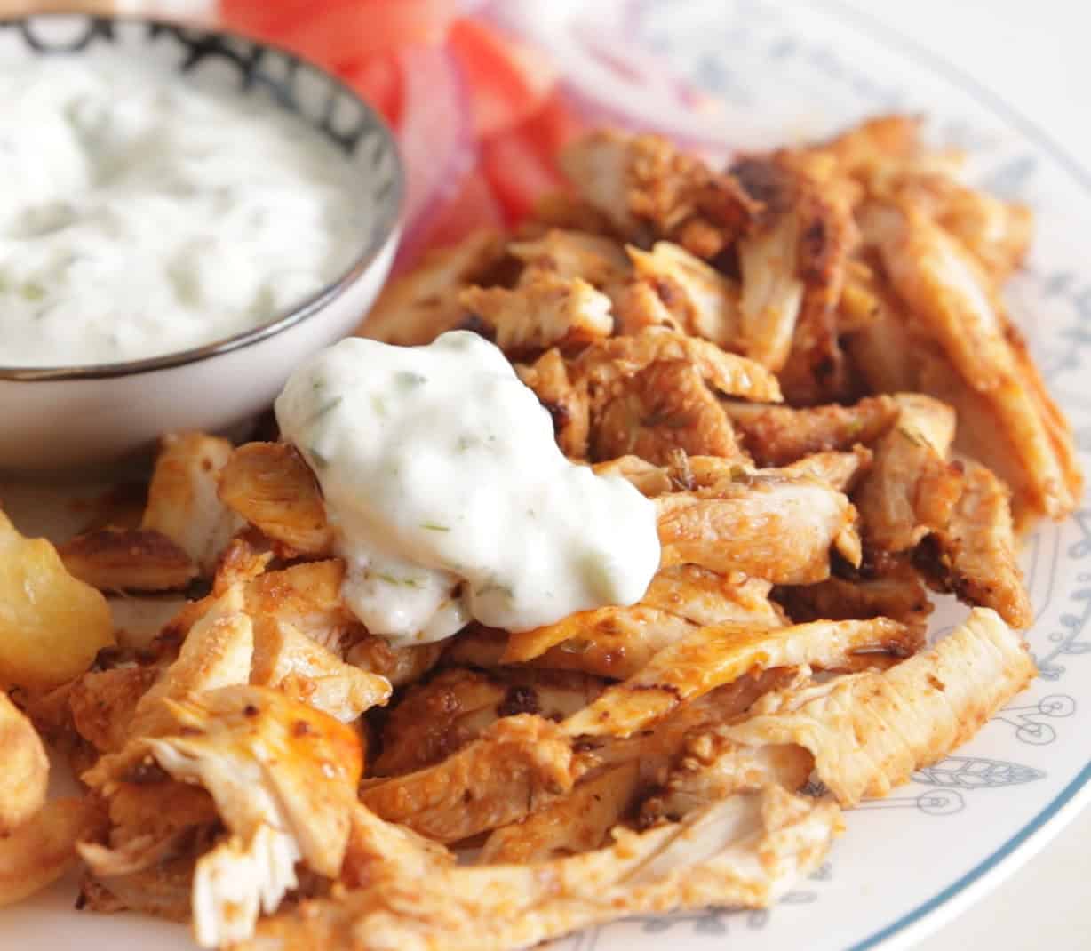A plate of seasoned, sliced chicken topped with a dollop of creamy white tzatziki sauce, served next to a bowl of more sauce and fresh vegetables.