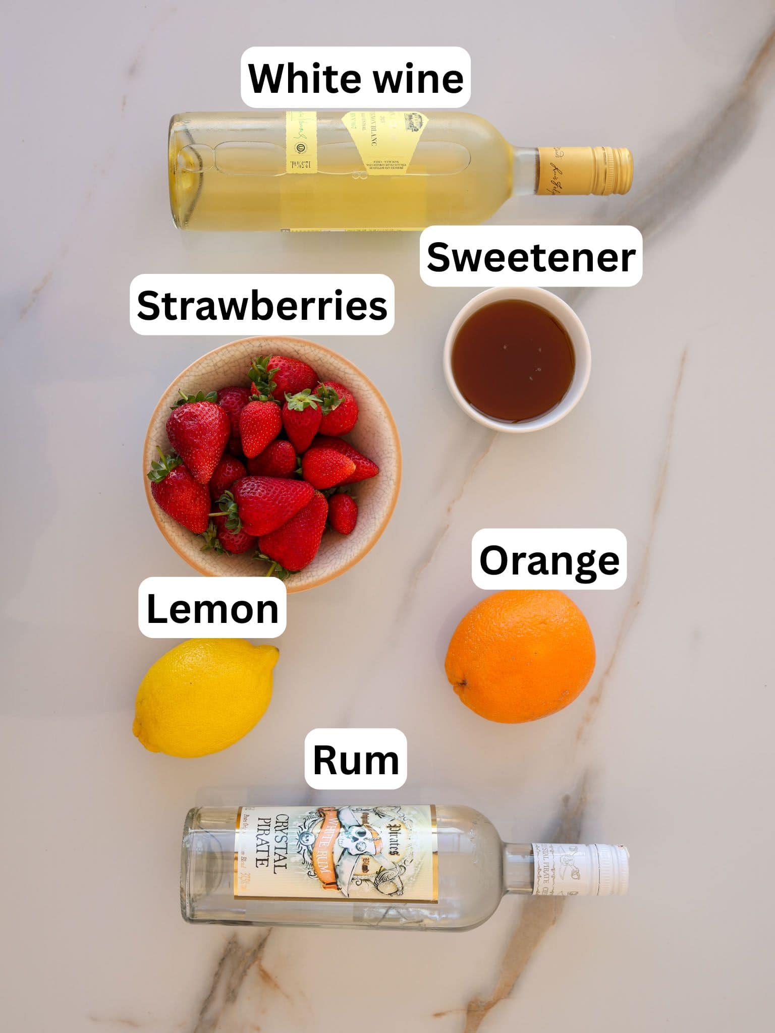 A flat lay of ingredients for a drink on a marble surface: a bottle of white wine, a bowl of strawberries, a lemon, an orange, a small bowl of sweetener, and a bottle of rum, each labeled.