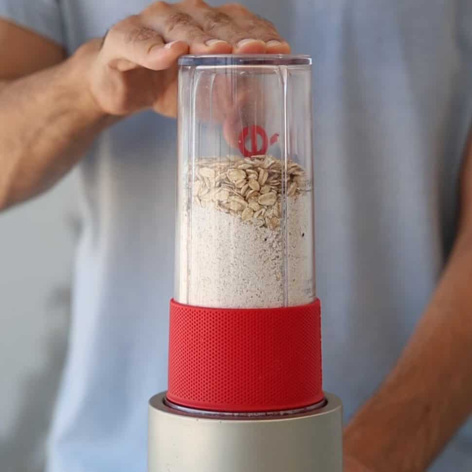A person holds a blender with a clear container filled with oats and oat flour, ready to blend, against a blurred background.
