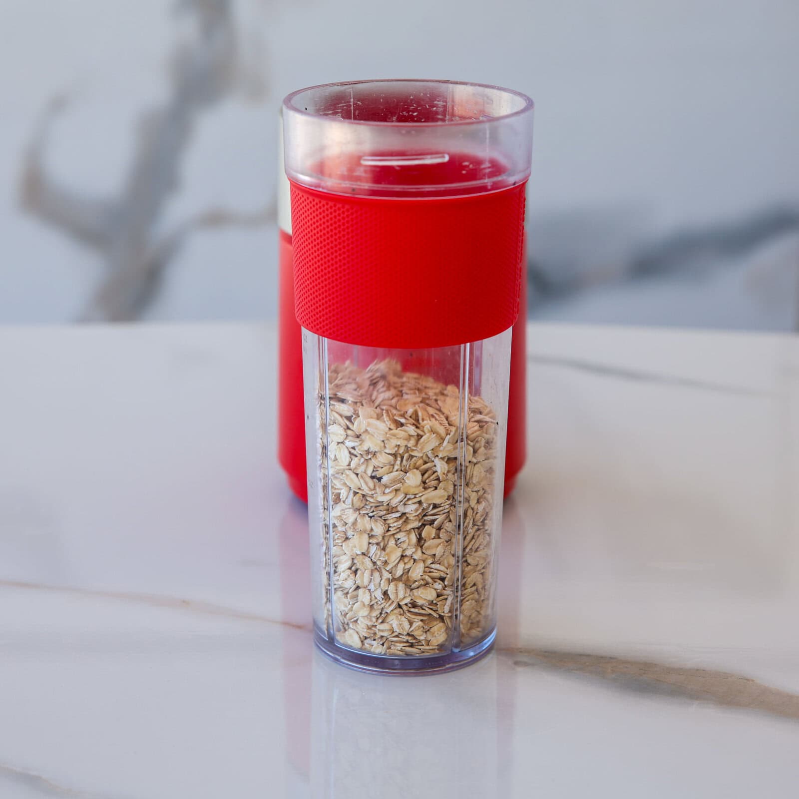 A clear tumbler with a red lid and red grip, filled halfway with dry rolled oats, sits on a white marble surface with a blurred background.