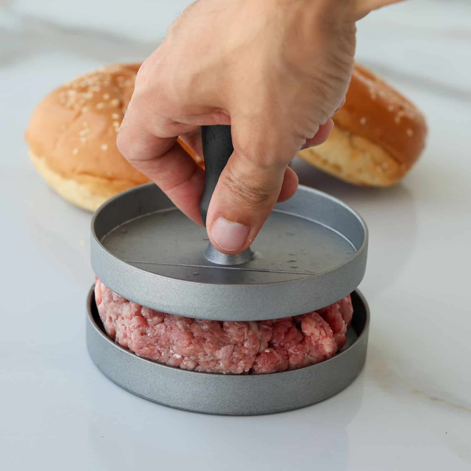 A hand presses raw ground meat into a patty using a metal burger press, with two sesame seed hamburger buns in the background on a white surface.
