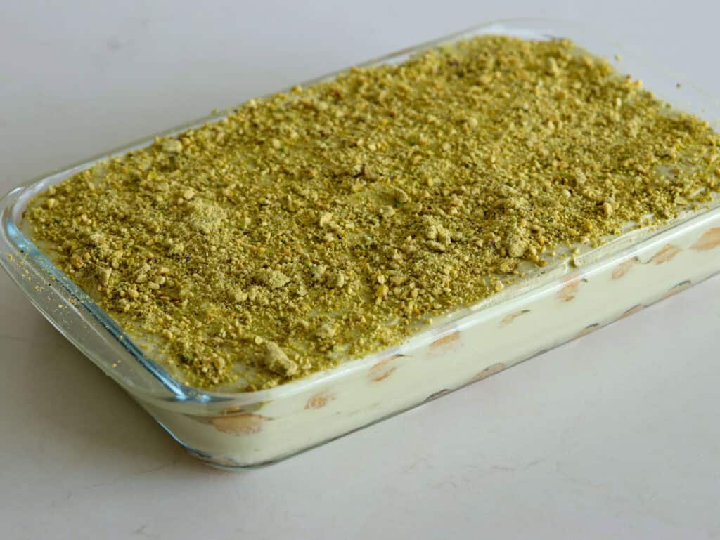 A rectangular glass dish filled with a pistachio dessert topped with a thick layer of finely crushed pistachios, showing creamy layers and biscuit pieces along the sides.