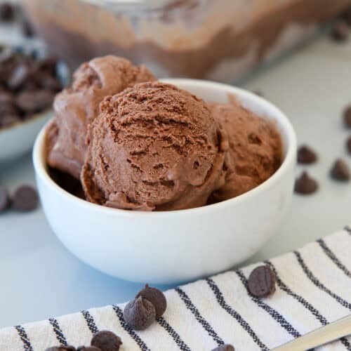 A white bowl filled with scoops of chocolate ice cream sits on a striped cloth, surrounded by scattered chocolate chips and a bowl of chocolate chips in the background.
