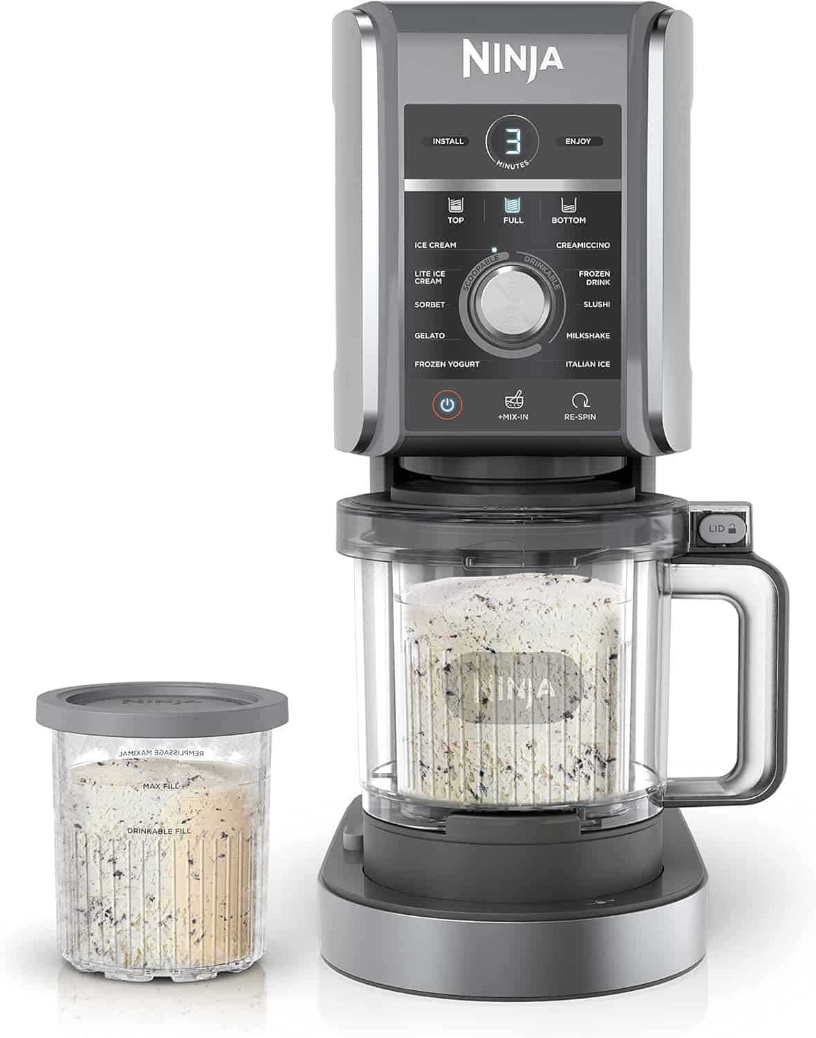 A Ninja ice cream maker with a clear blending container attached, filled with a creamy dessert, sits next to a matching storage container with a gray lid, both containing ice cream with cookie pieces.