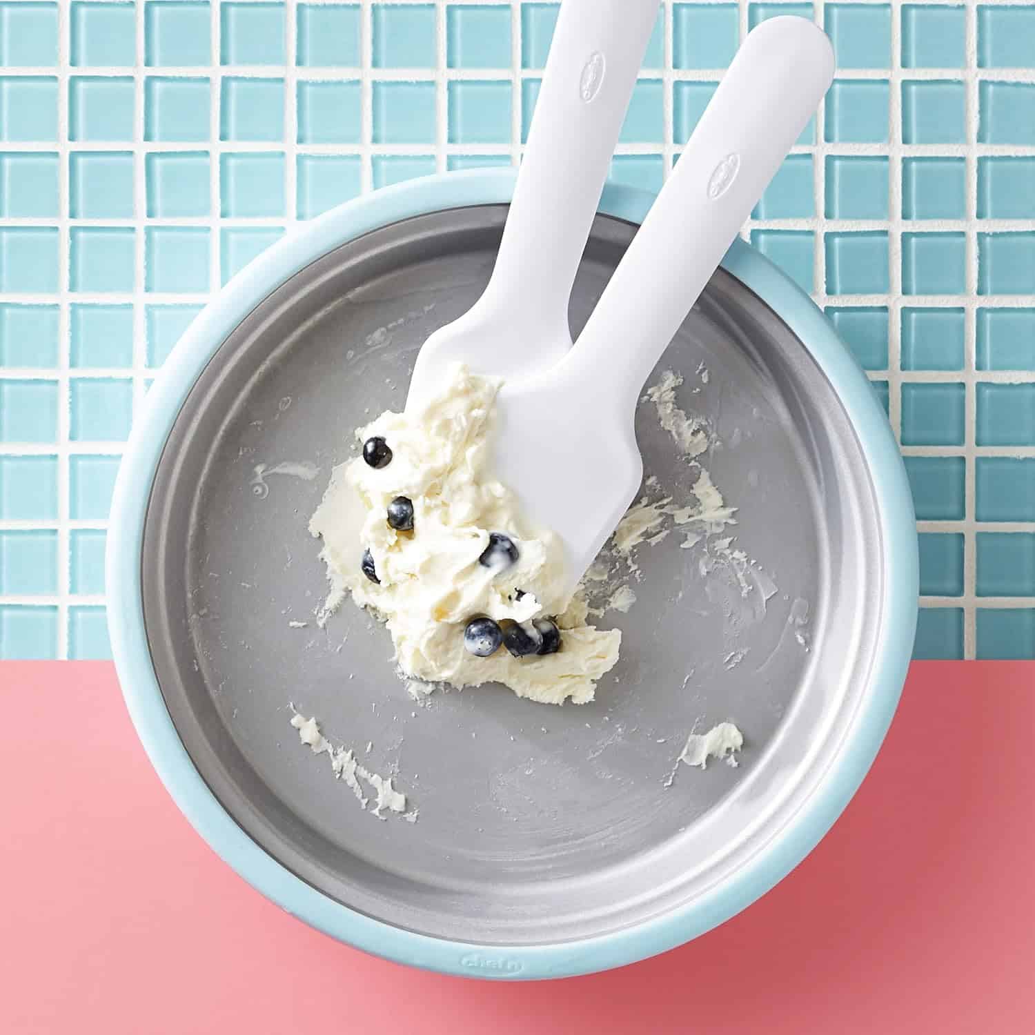 A metal pan with a small amount of creamy ice cream and blueberries, being scraped by two white spatulas, sits on a blue and pink tiled surface.