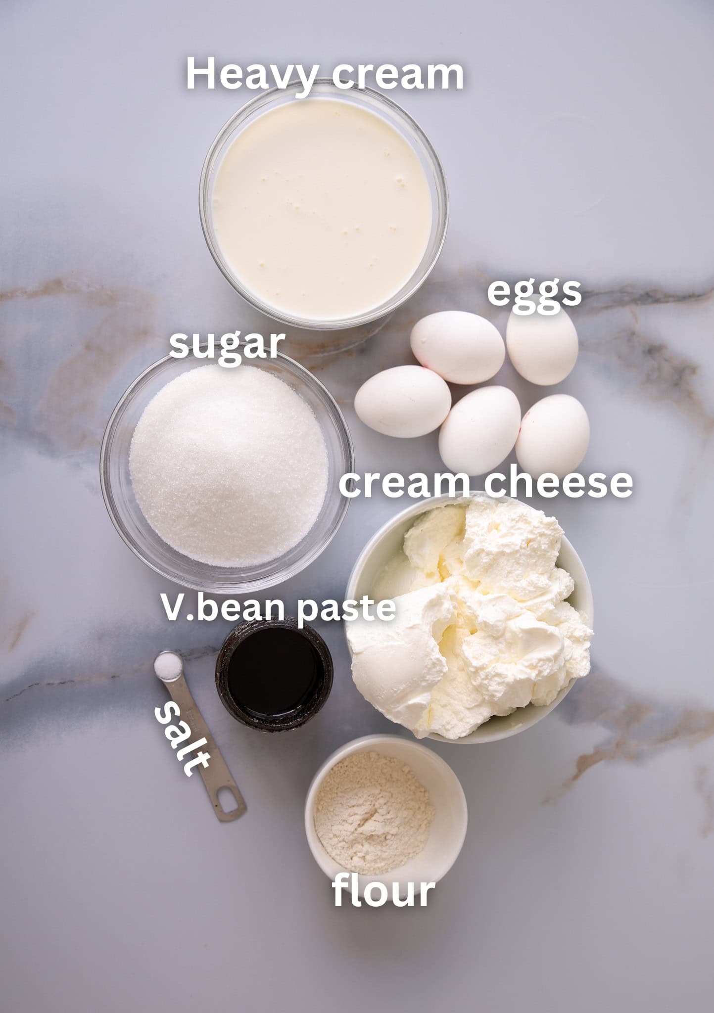 Top-down view of baking ingredients on a marble surface, including bowls of heavy cream, sugar, cream cheese, vanilla bean paste, flour, salt, and six whole eggs, each labeled with text.