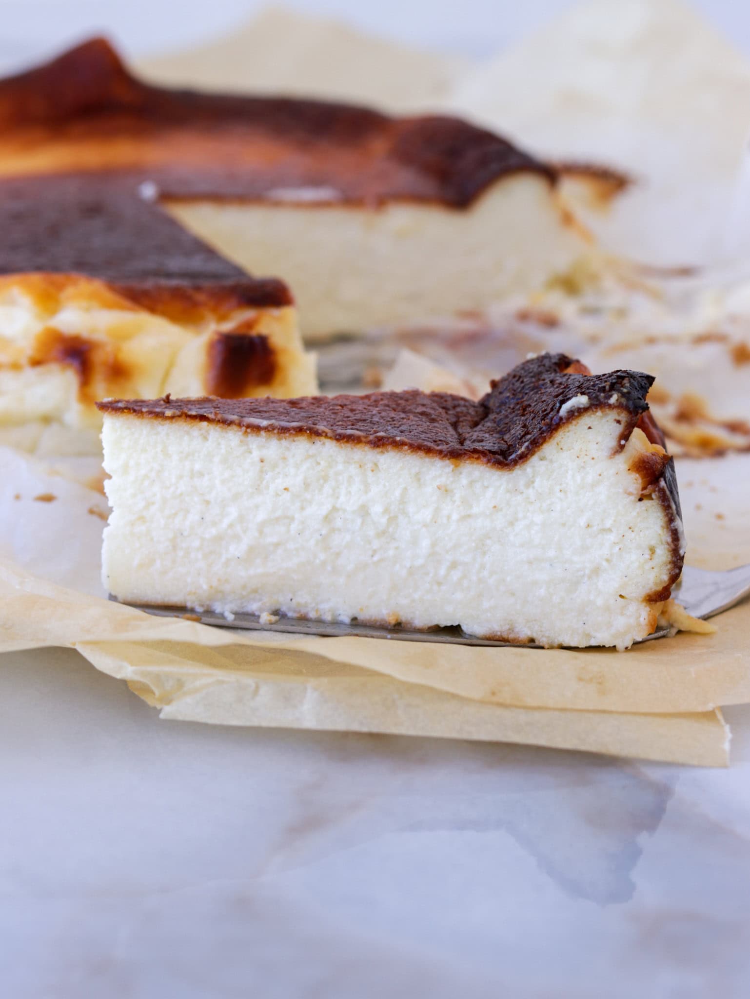 A slice of Basque burnt cheesecake with a dark, caramelized top sits on parchment paper, with the remaining cheesecake and another slice visible in the background.