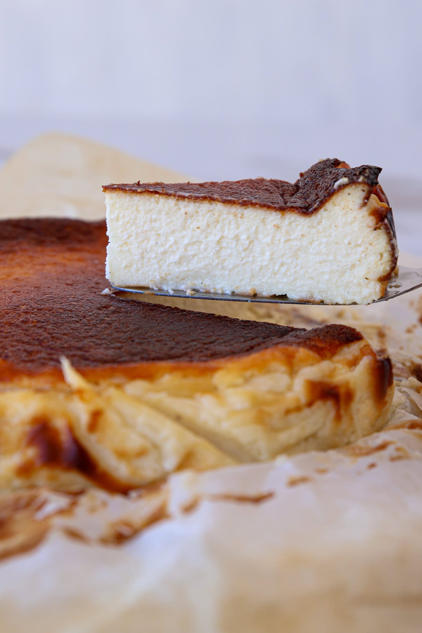 A close-up of a creamy slice of Basque burnt cheesecake being lifted from the whole cake, which sits on parchment paper. The cheesecake has a dark, caramelized top and a light, fluffy interior.