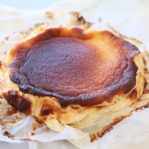A rustic Basque burnt cheesecake with a caramelized, dark brown top sits on crumpled parchment paper, its edges uneven and slightly cracked. The cheesecake’s creamy interior is visible around the sides.